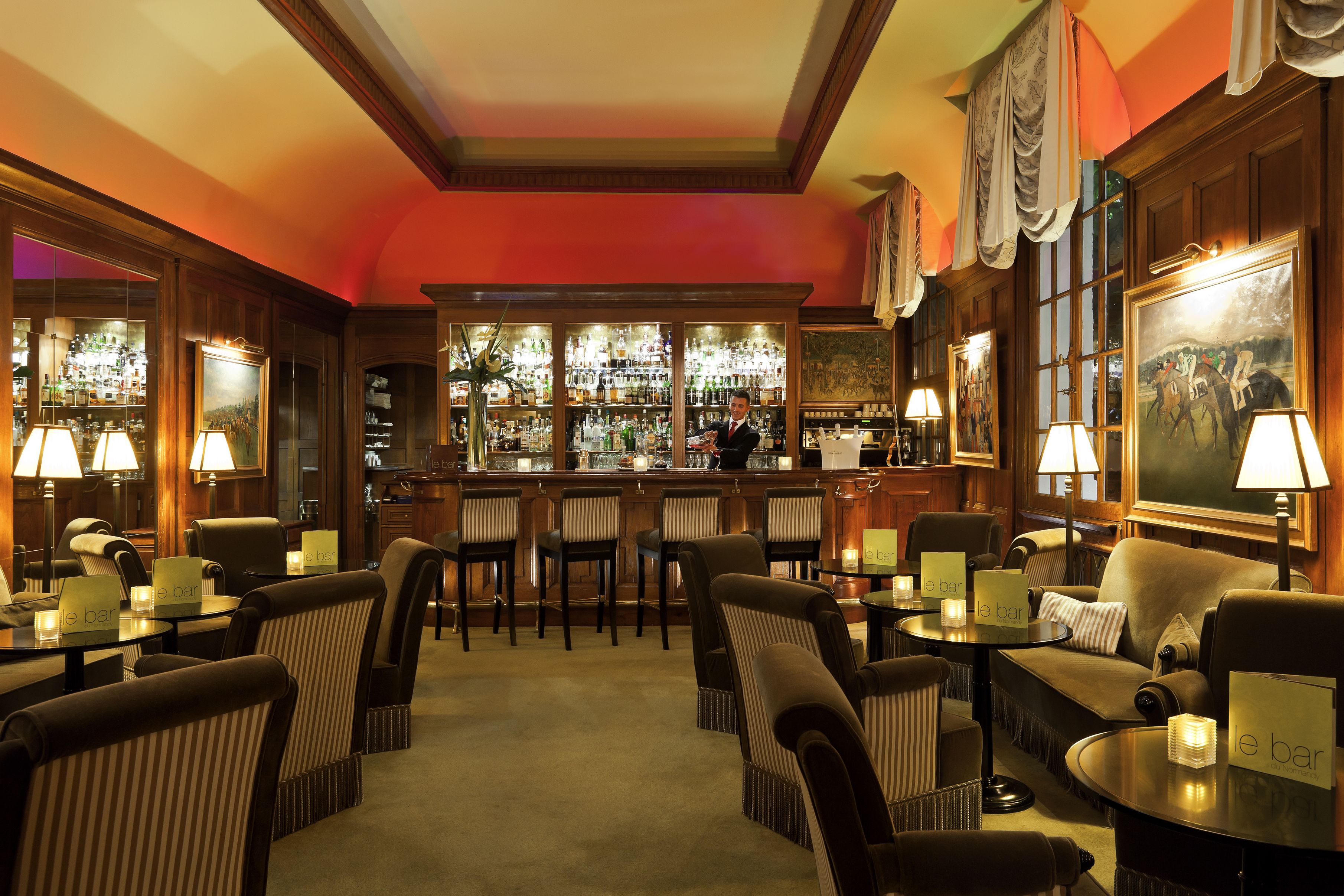 A bar area with multiple seating options, including bar stools and cushioned chairs, with a bar counter and shelves stocked with bottles.