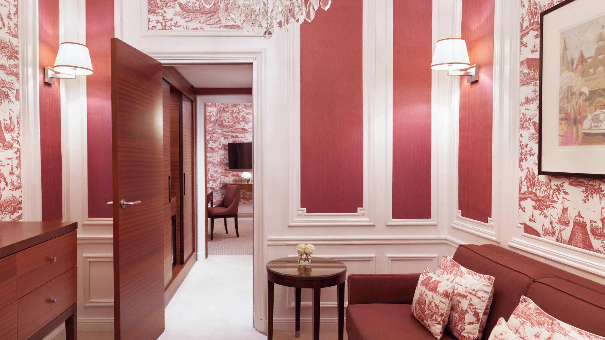 A hallway with a wooden door, a round table with a vase, and a red sofa with patterned cushions.