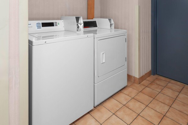 A front view of a white front-loading washer and dryer unit with a control panel, located in a room with tiled flooring and a door to the right.