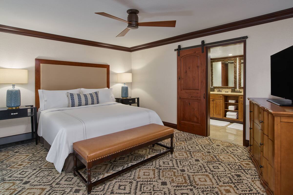 Habitación de hotel con cama, dos lámparas de noche, una banca, ventilador de techo, armario de madera y baño visible a través de una puerta abierta.