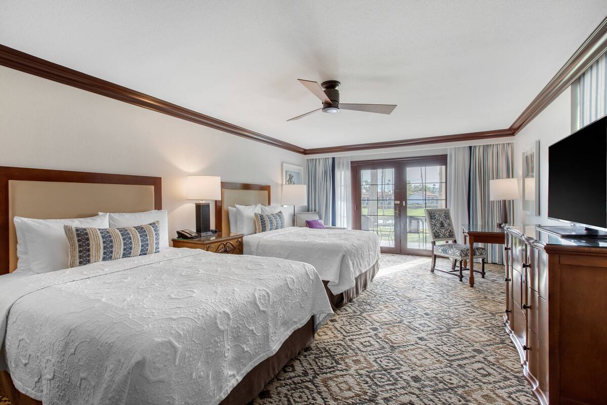 Habitación de hotel con dos camas, ventilador de techo, televisión, ventana grande con cortinas y vista al exterior.