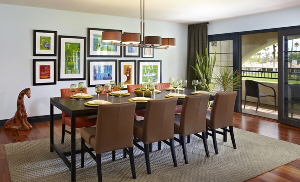 Comedor con una gran mesa dispuesta para seis personas, rodeada de sillas marrones. La habitación tiene un amplio ventanal con vista a palmeras y un balcón.