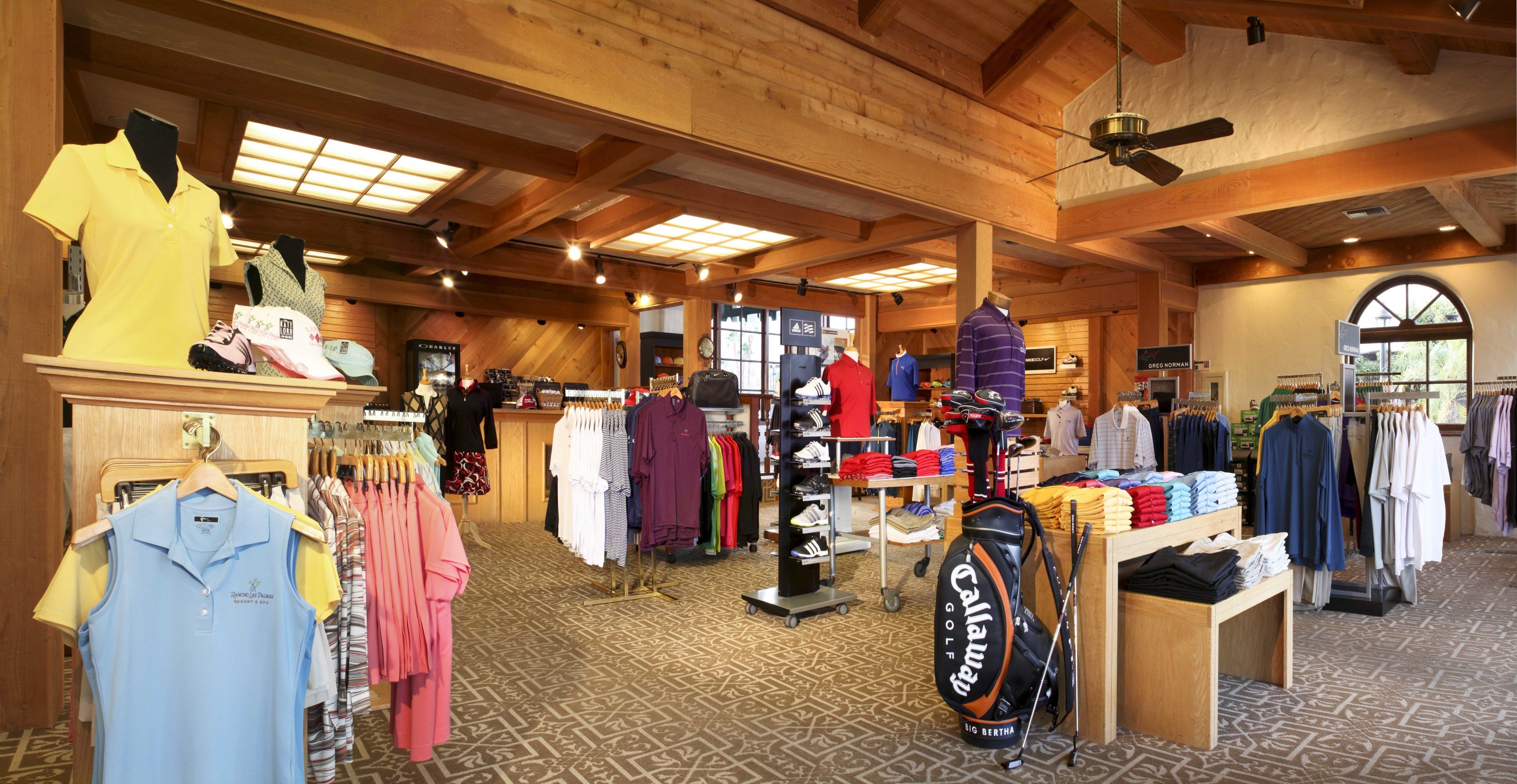 A clothing store with racks of shirts and pants, a golf bag display, and a ceiling fan.