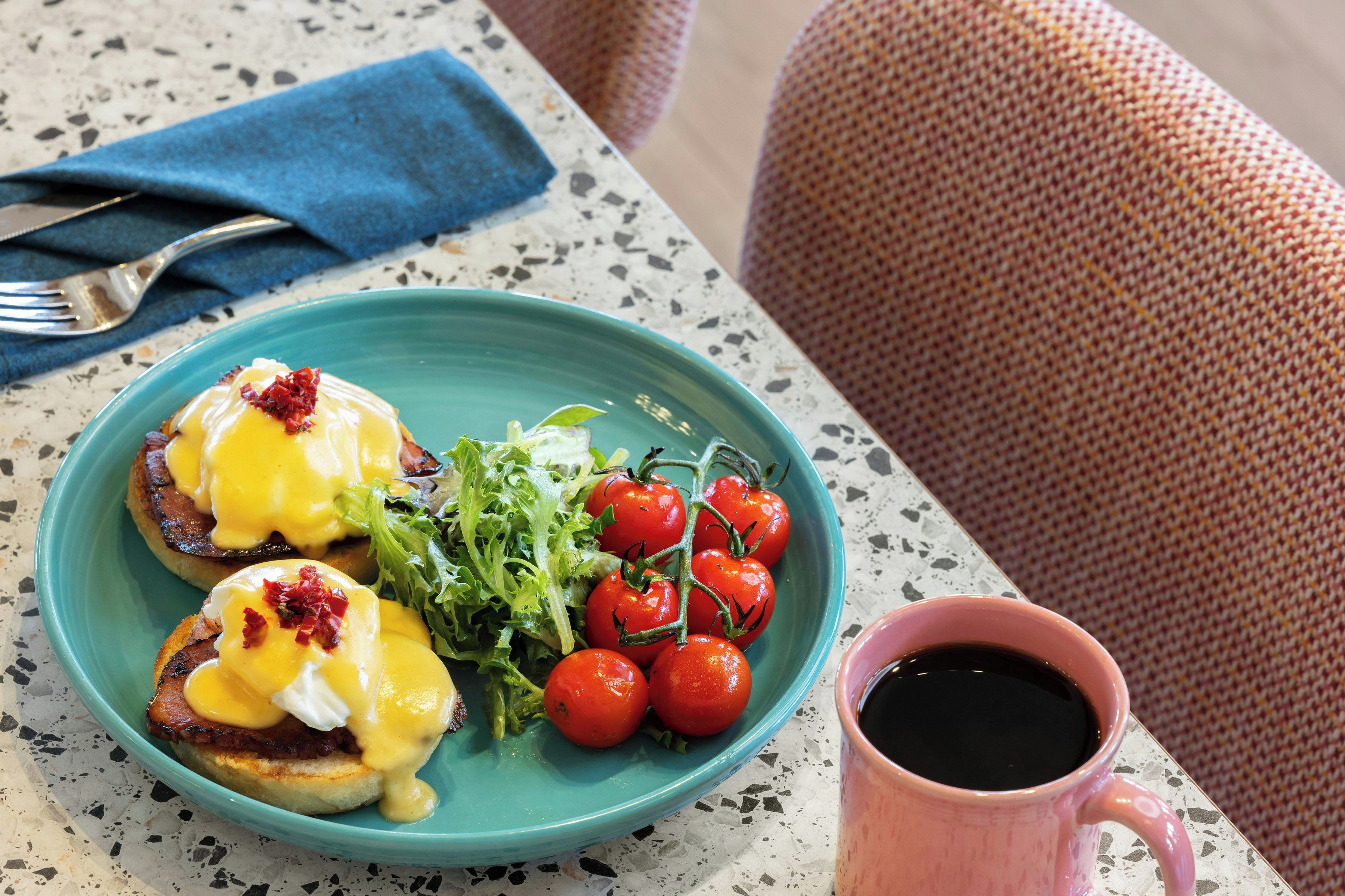 En tallerken med eggs benedict og kirsebærtomater, en kop kaffe og en blå serviet på et plettet bord.