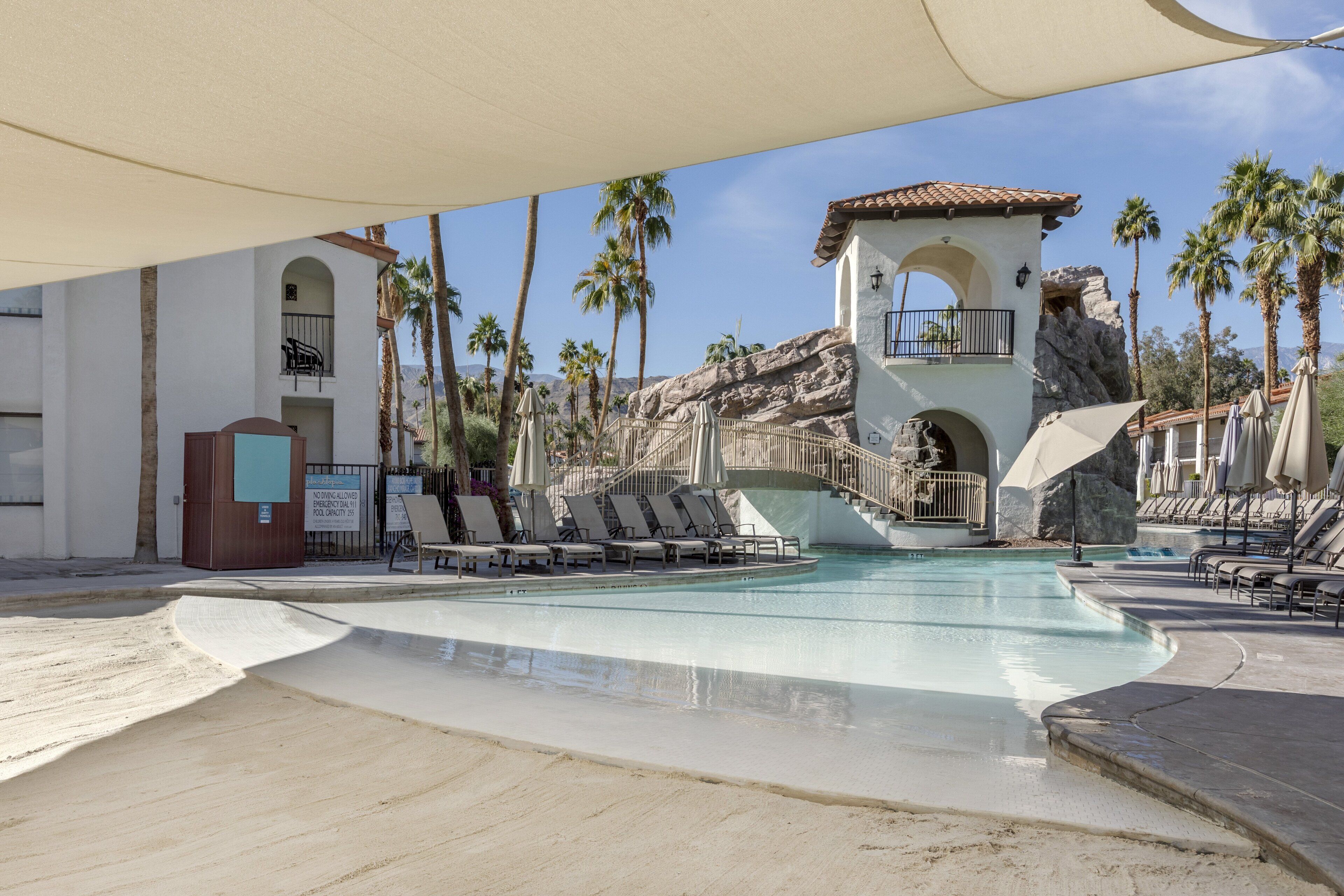 A pool area with lounge chairs, palm trees, and a building with a rock facade.