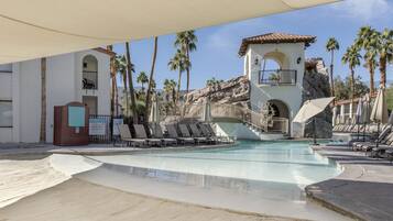 A pool area with lounge chairs, palm trees, and a building with a rock facade.