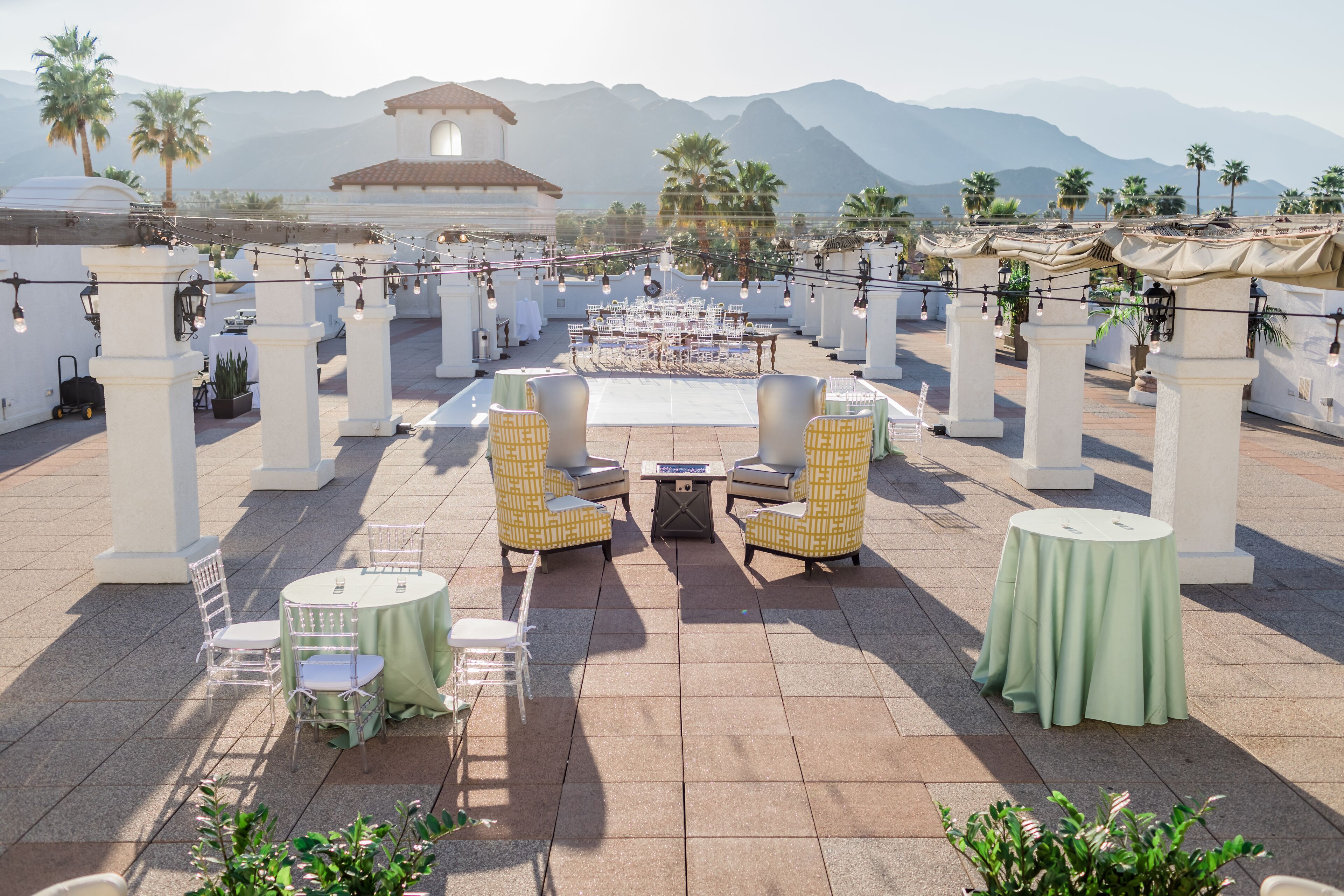 An outdoor patio with round tables and chairs, palm trees, and a mountain backdrop.