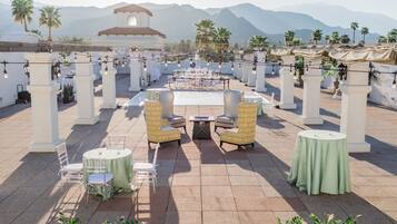 An outdoor patio with round tables and chairs, palm trees, and a mountain backdrop.