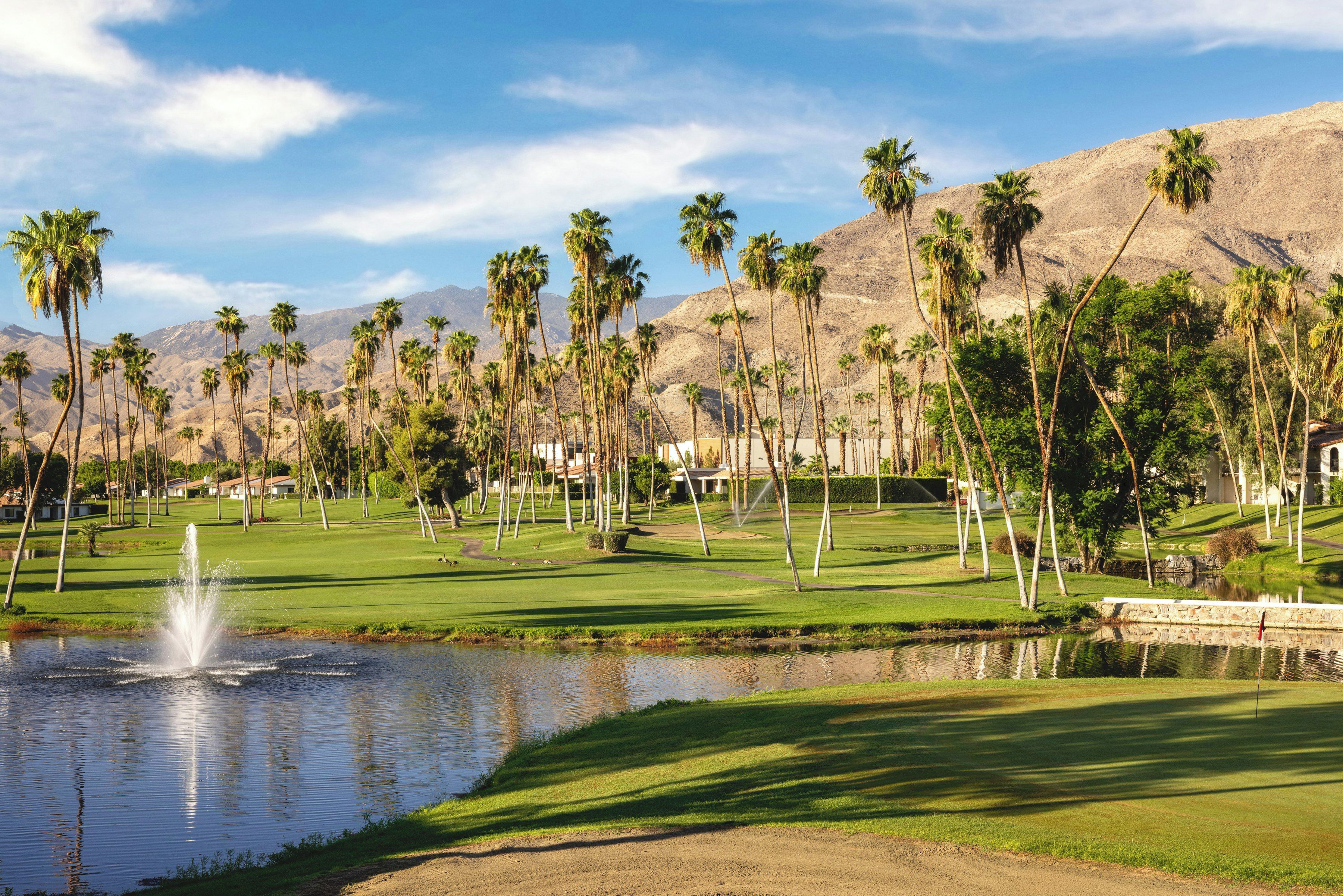 Un campo de golf con un estanque, palmeras y montañas al fondo.