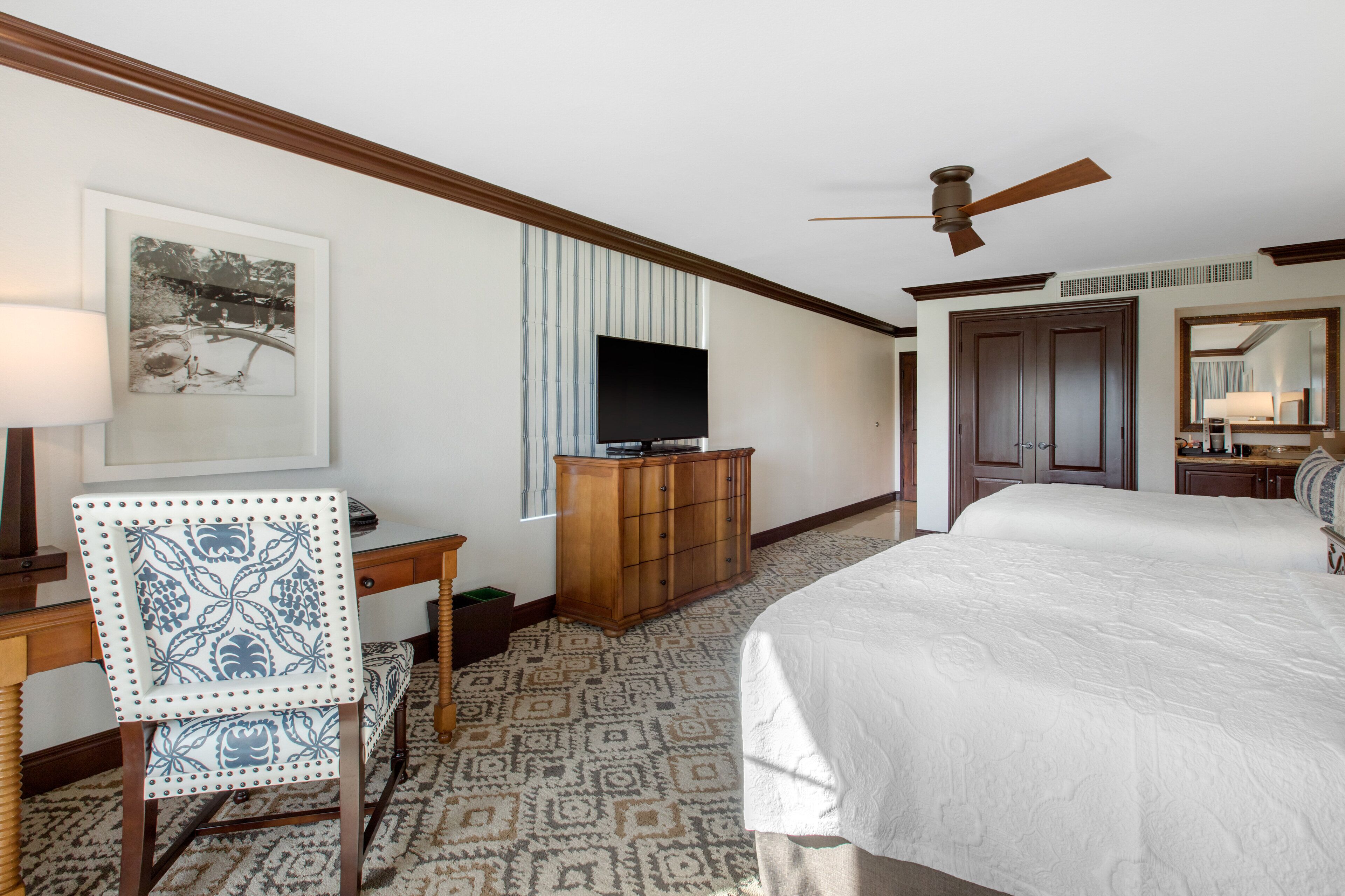 A hotel room with a bed, a desk with a chair, a television, a wardrobe, and a ceiling fan.