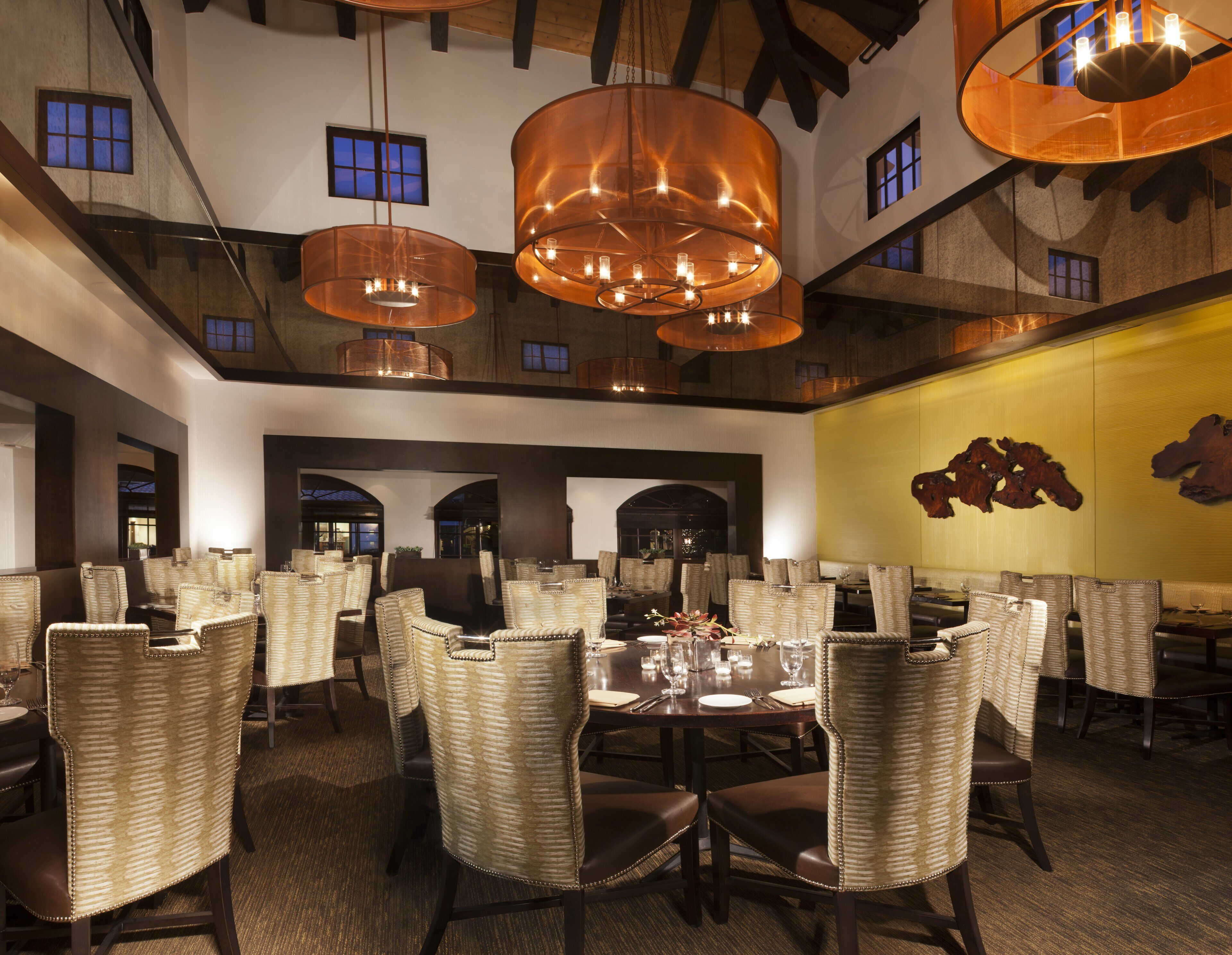 A spacious dining area with high ceilings and multiple hanging copper chandeliers. Tables are set with chairs, and the walls feature large windows and a decorative wall piece.