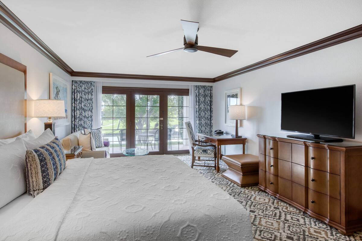 Habitación de hotel con una cama grande, un escritorio con silla, televisor y balcón con vista a árboles.