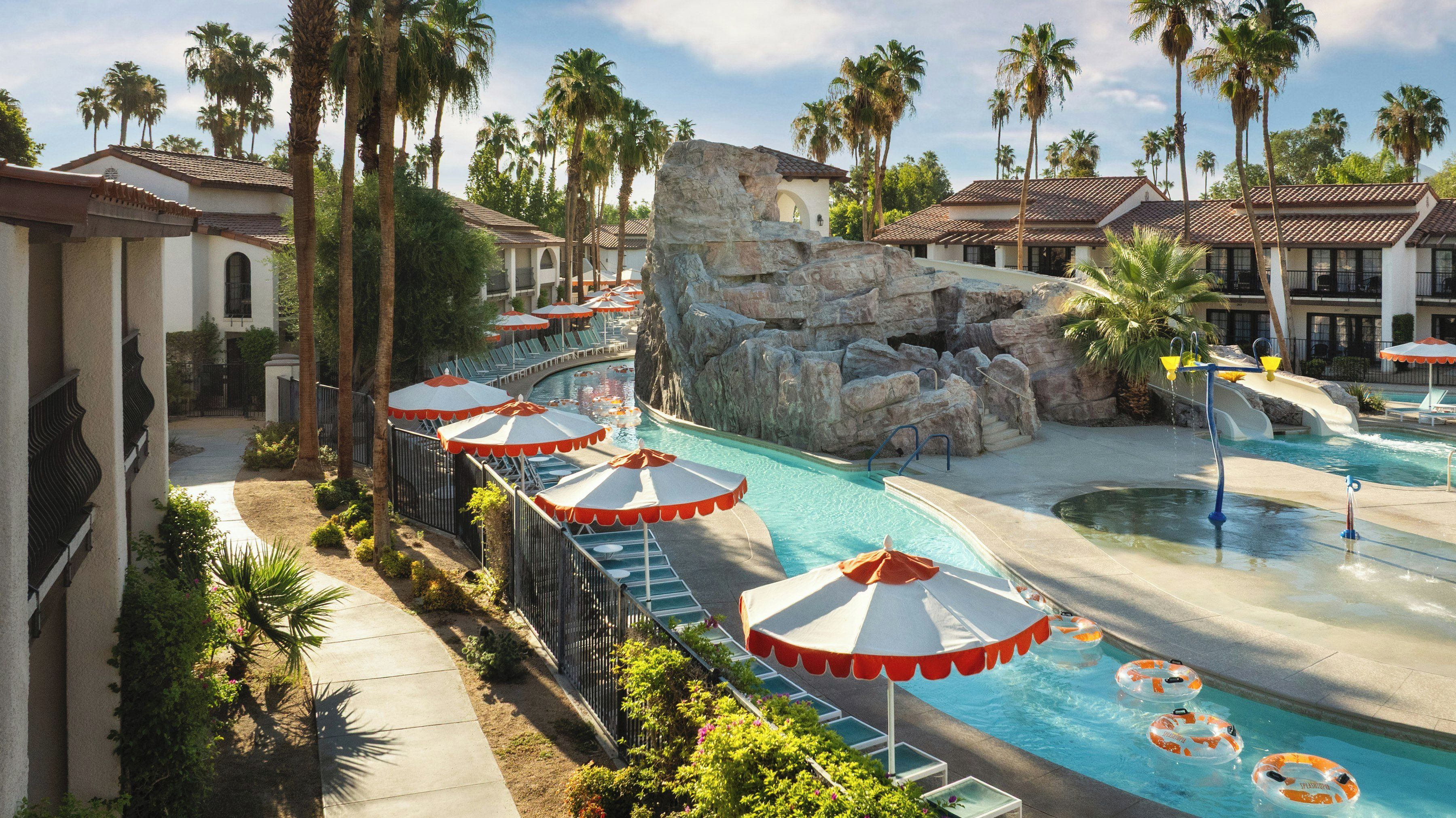 A resort pool area with multiple lounge chairs and umbrellas, a rock formation, and palm trees.