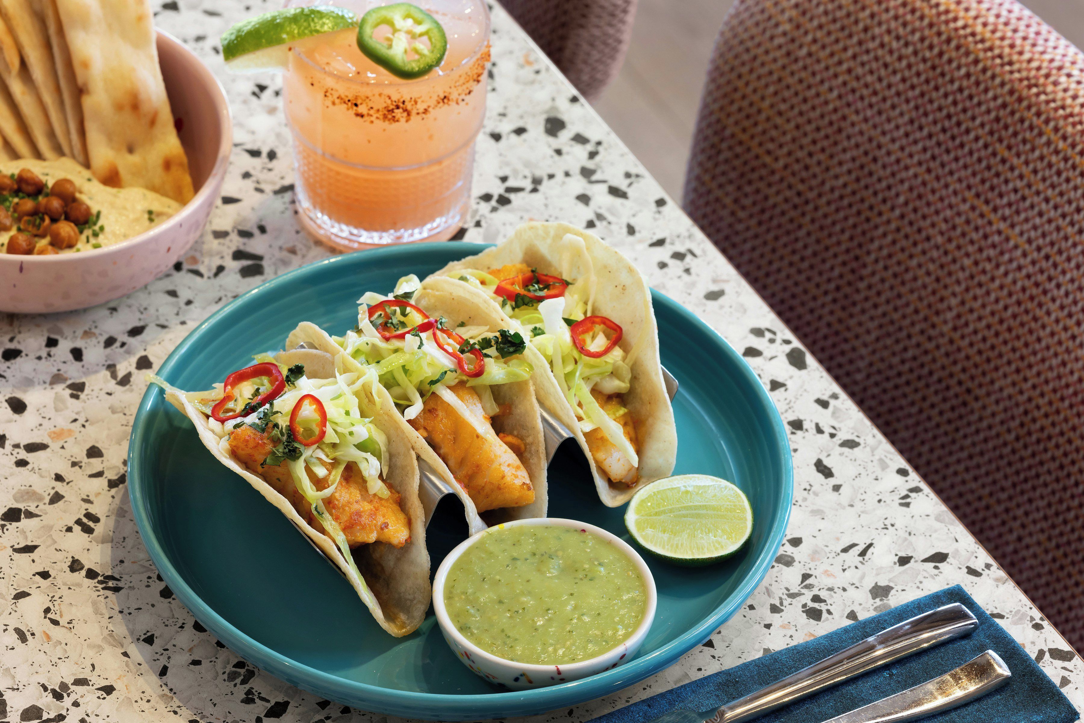 A plate of fish tacos with a side of green sauce and a lime wedge, accompanied by a glass of a pink beverage and a bowl of chips.