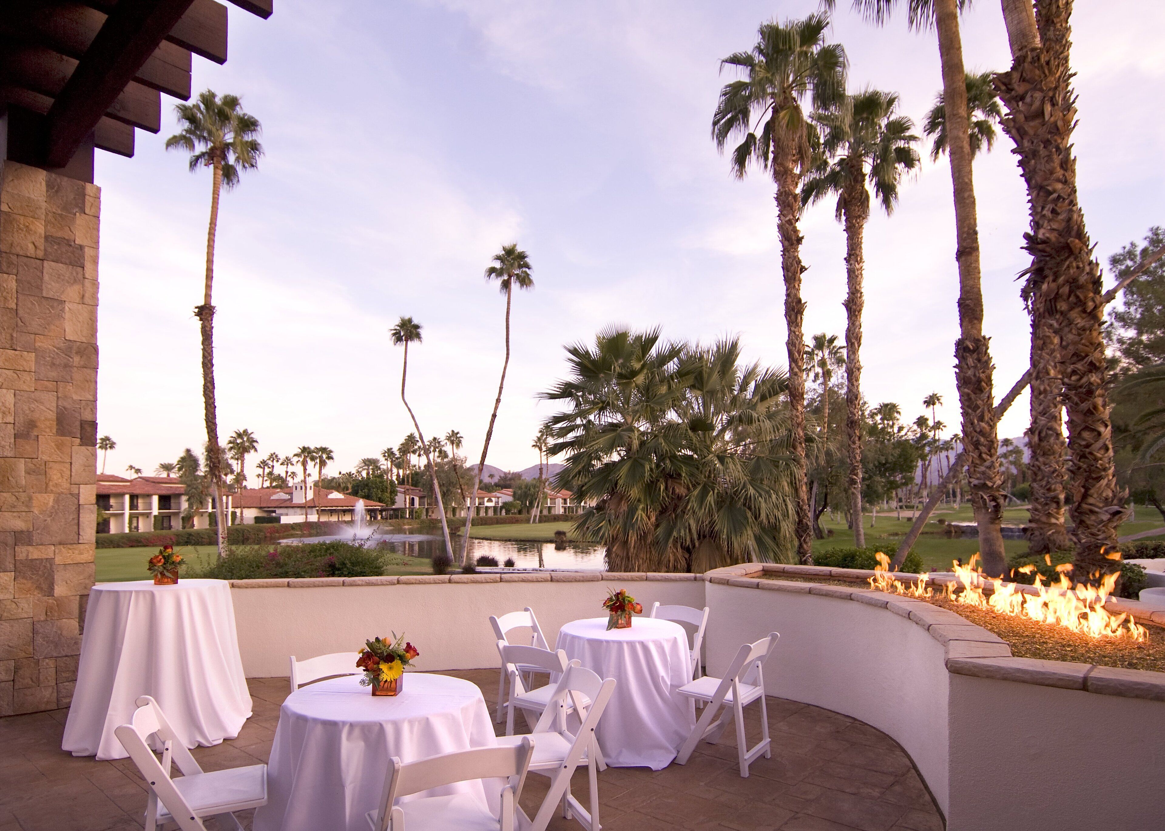 An outdoor patio with round tables and white chairs, surrounded by palm trees and a view of a golf course.