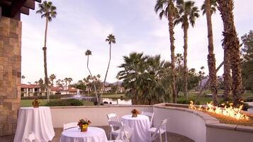 An outdoor patio with round tables and white chairs, surrounded by palm trees and a view of a golf course.