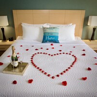 A neatly made bed with a heart-shaped decoration of rose petals and red heart-shaped objects, a bottle of champagne, and a pillow with the text "Beach is my Happy place".