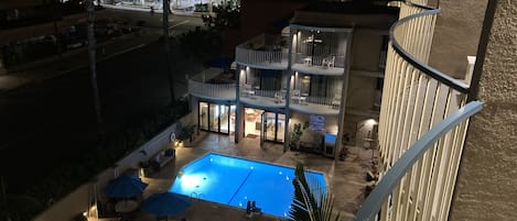 A rooftop pool area with multiple lounge chairs and umbrellas, surrounded by a multi-story building with balconies and outdoor seating.