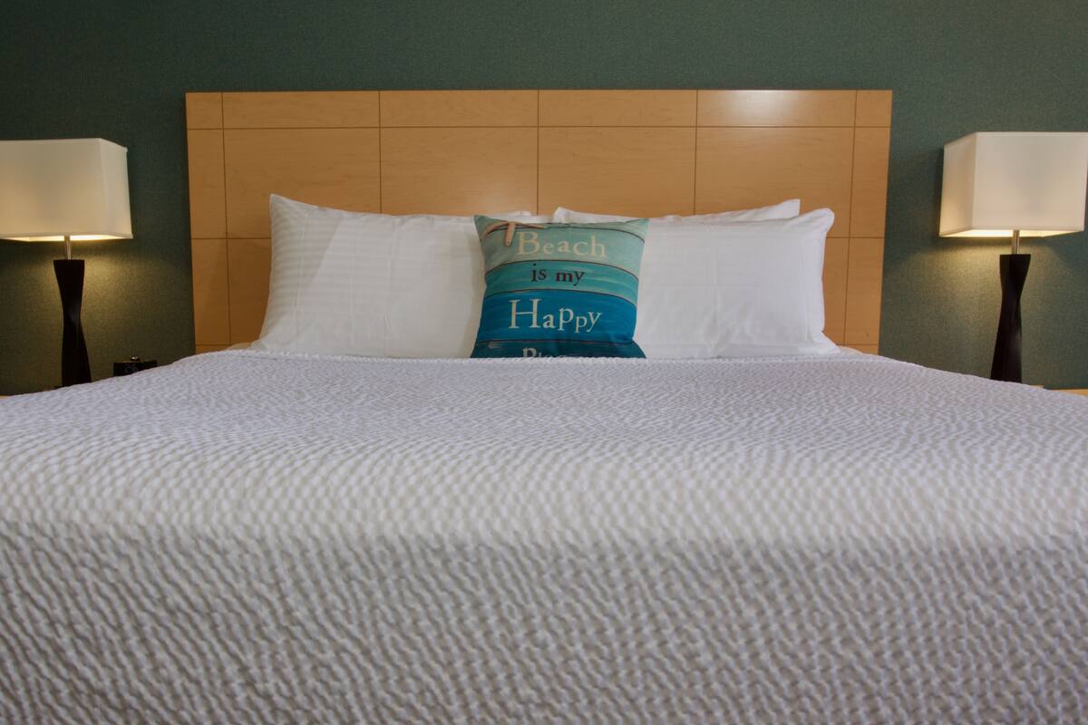 Un lit bien fait, recouvert d’une couette blanche et agrémenté de deux oreillers, dont l’un est orné d’une taie d’oreiller sur le thème de la plage. Deux lampes de chevet sont placées de part et d’autre du lit.
