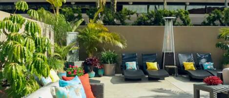 An outdoor seating area with lounge chairs and cushions, surrounded by potted plants and a modern building in the background.
