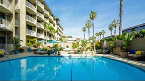 A swimming pool with lounge chairs and umbrellas, surrounded by palm trees and a multi-story building with balconies. - Inn By The Sea La Jolla (La Jolla)