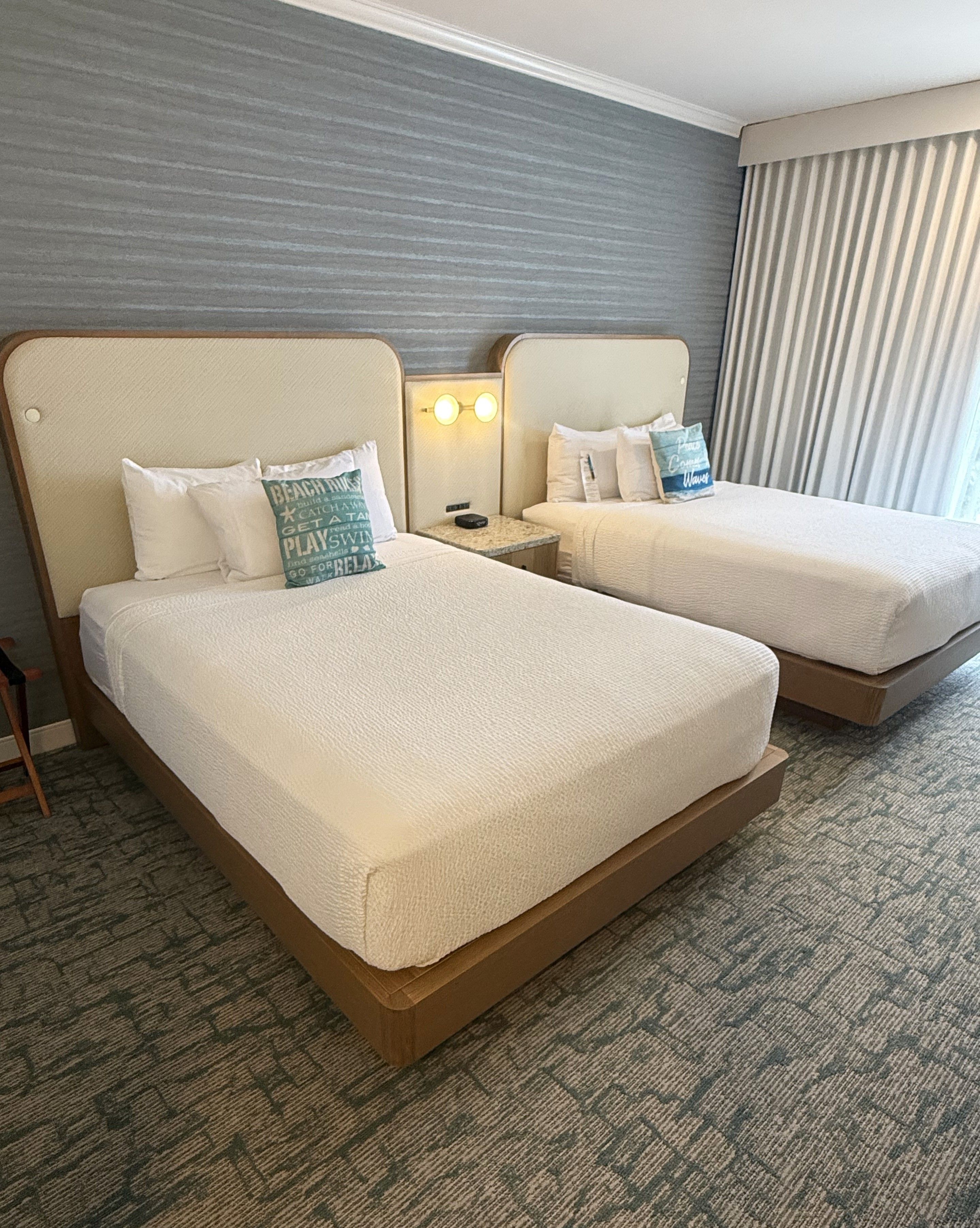 A hotel room with two beds, a nightstand, a lamp, and a window with curtains.