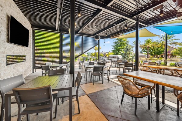 Un café en plein air avec des tables et des chaises, un mur en briques et une grande fenêtre avec des stores.
