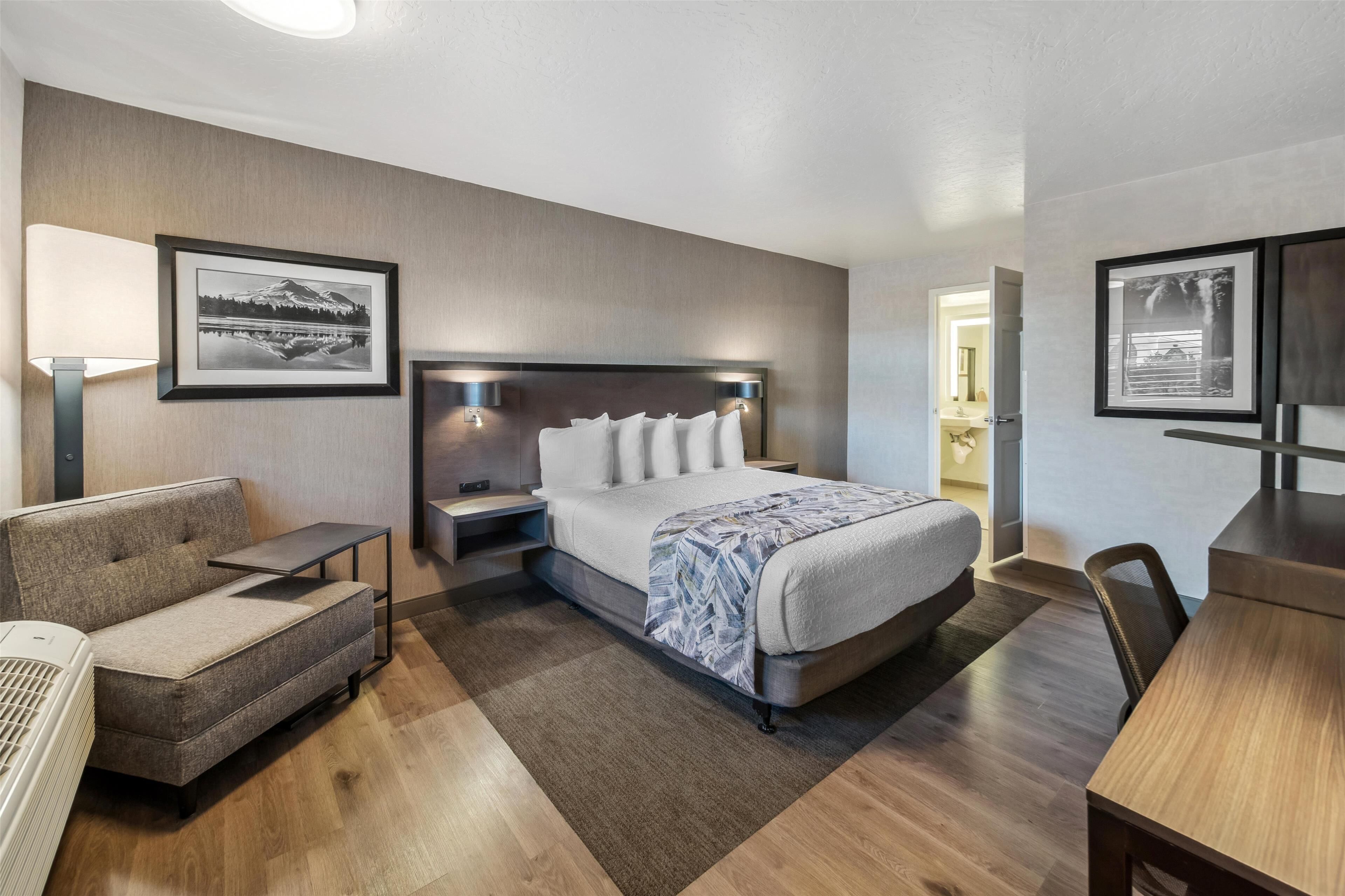 Een moderne hotelkamer met een groot bed, een bank, een bureau en een stoel. Er hangen ingelijste foto’s aan de muur en op het nachtkastje staat een lamp.