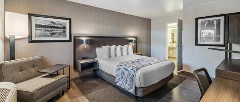 A modern hotel room with a large bed, a sofa, a desk, and a chair. There are framed pictures on the walls and a lamp on the nightstand.