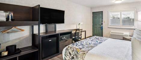 A hotel room with a bed, a desk with a television, a small refrigerator, a chair, a shelf with a hanging garment, and a window with blinds.