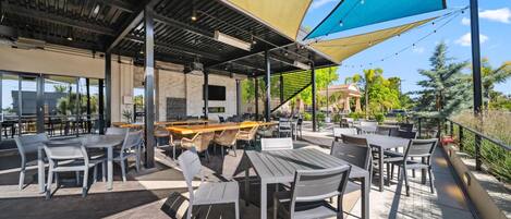 Un espace extérieur aménagé en terrasse, avec des tables et des chaises, protégé par de grands auvents, et un coin bar avec des tabourets.