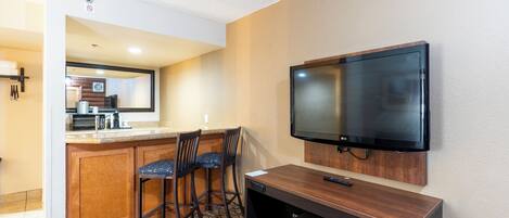 A compact hotel room with a kitchenette, a flat-screen TV mounted on the wall, a small desk, and two bar stools.