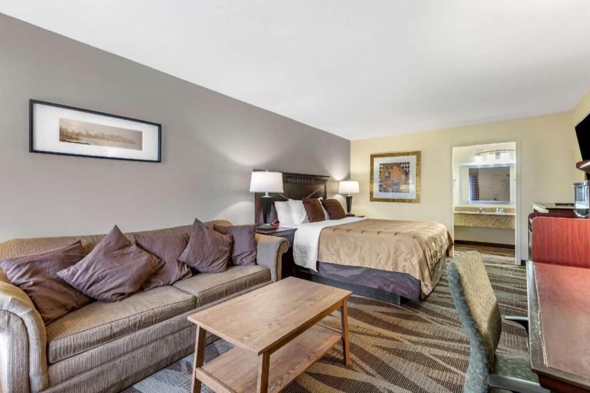 A hotel room with a bed, a sofa, a desk, a chair, a TV, and a bathroom visible through an open door.