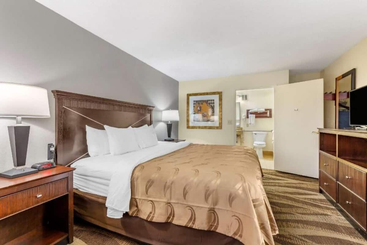 A hotel room with a bed, bedside tables, a television, a mirror, and a bathroom visible through an open door.