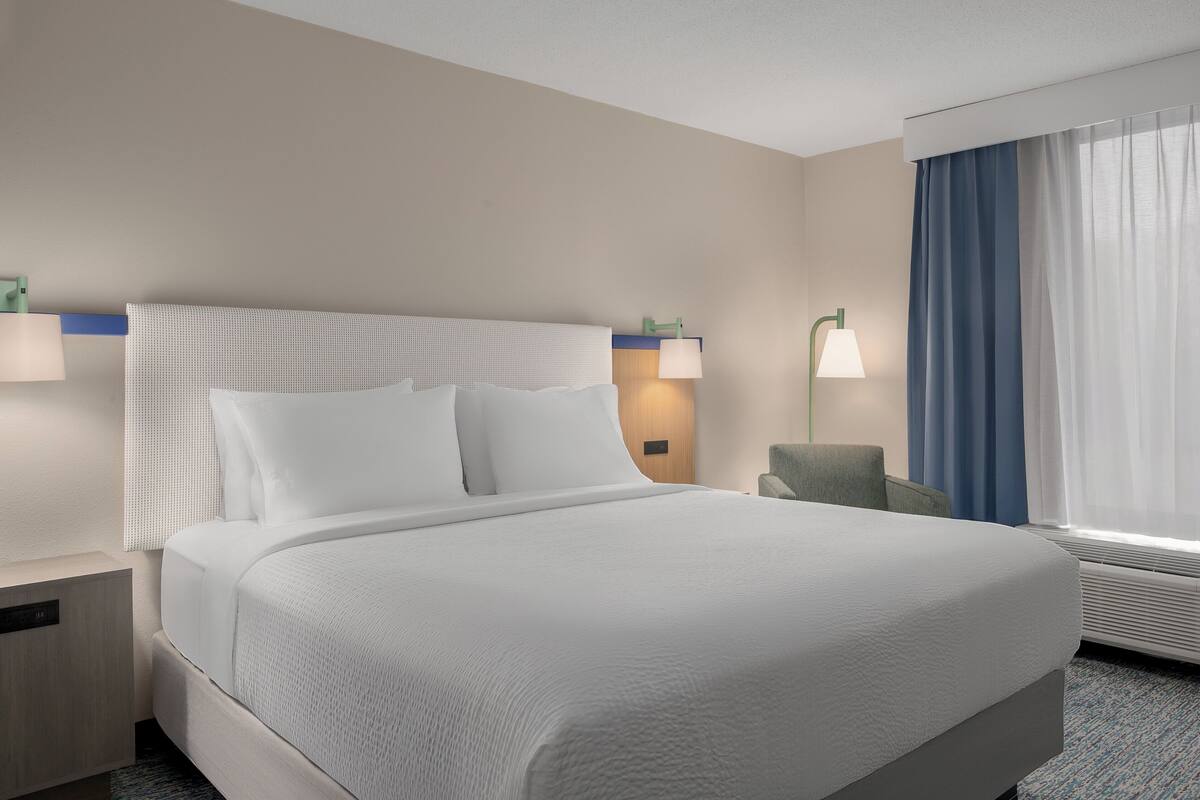 Une chambre d’hôtel avec un grand lit, deux tables de chevet, une chaise, une fenêtre avec des rideaux et une lampe fixée au mur.