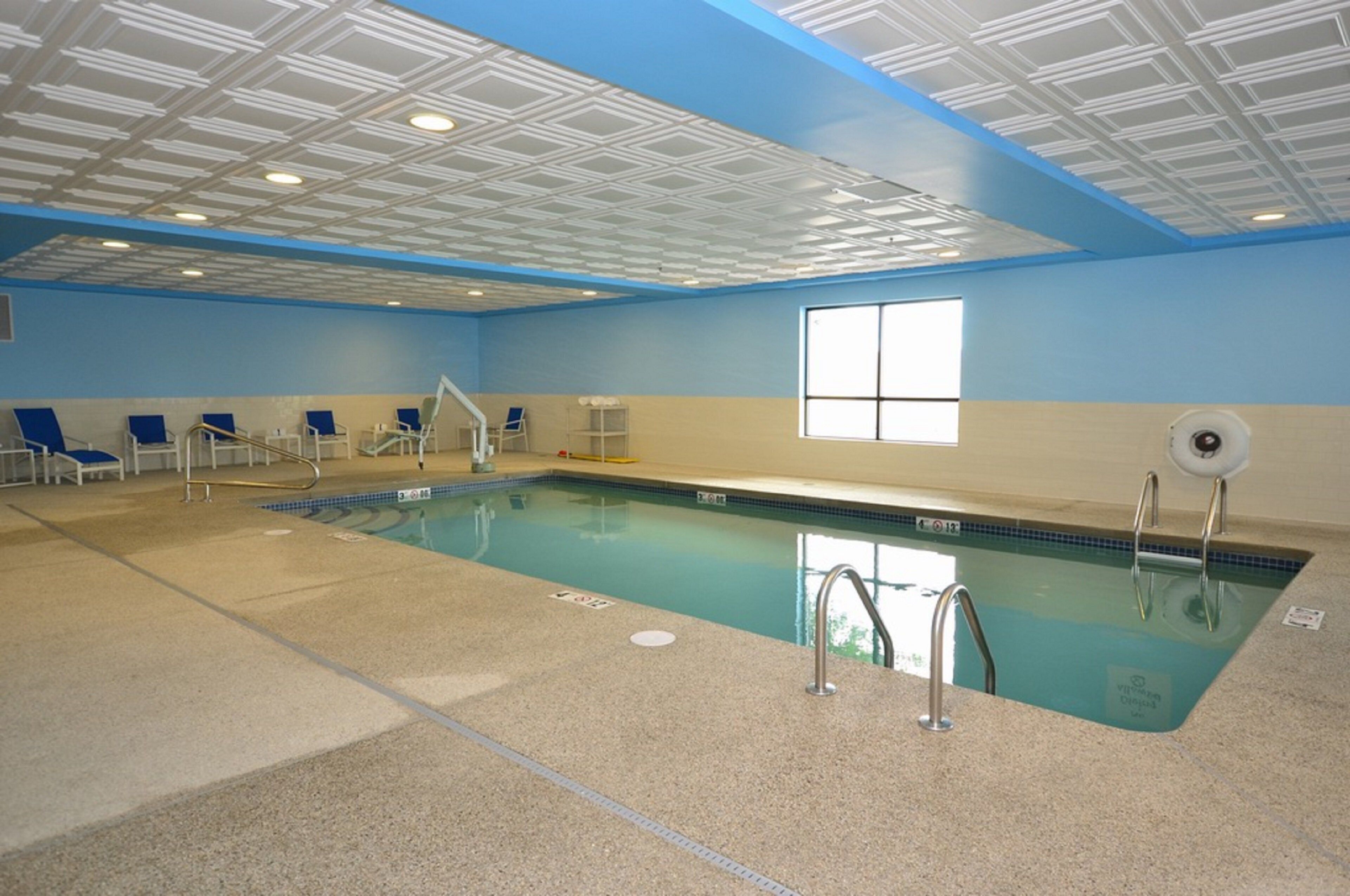 An indoor swimming pool with clear water, surrounded by blue chairs, a window, and a ceiling with a geometric pattern.