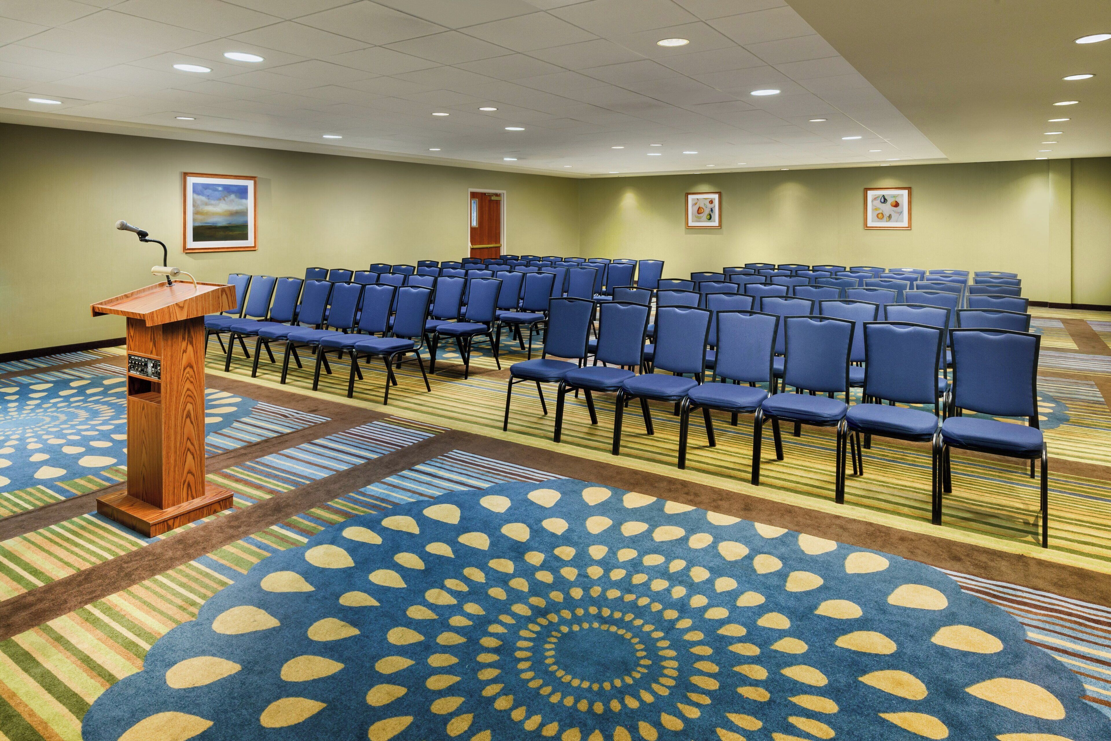 Un salón de conferencias con filas de sillas, un atril y una alfombra con diseño circular.