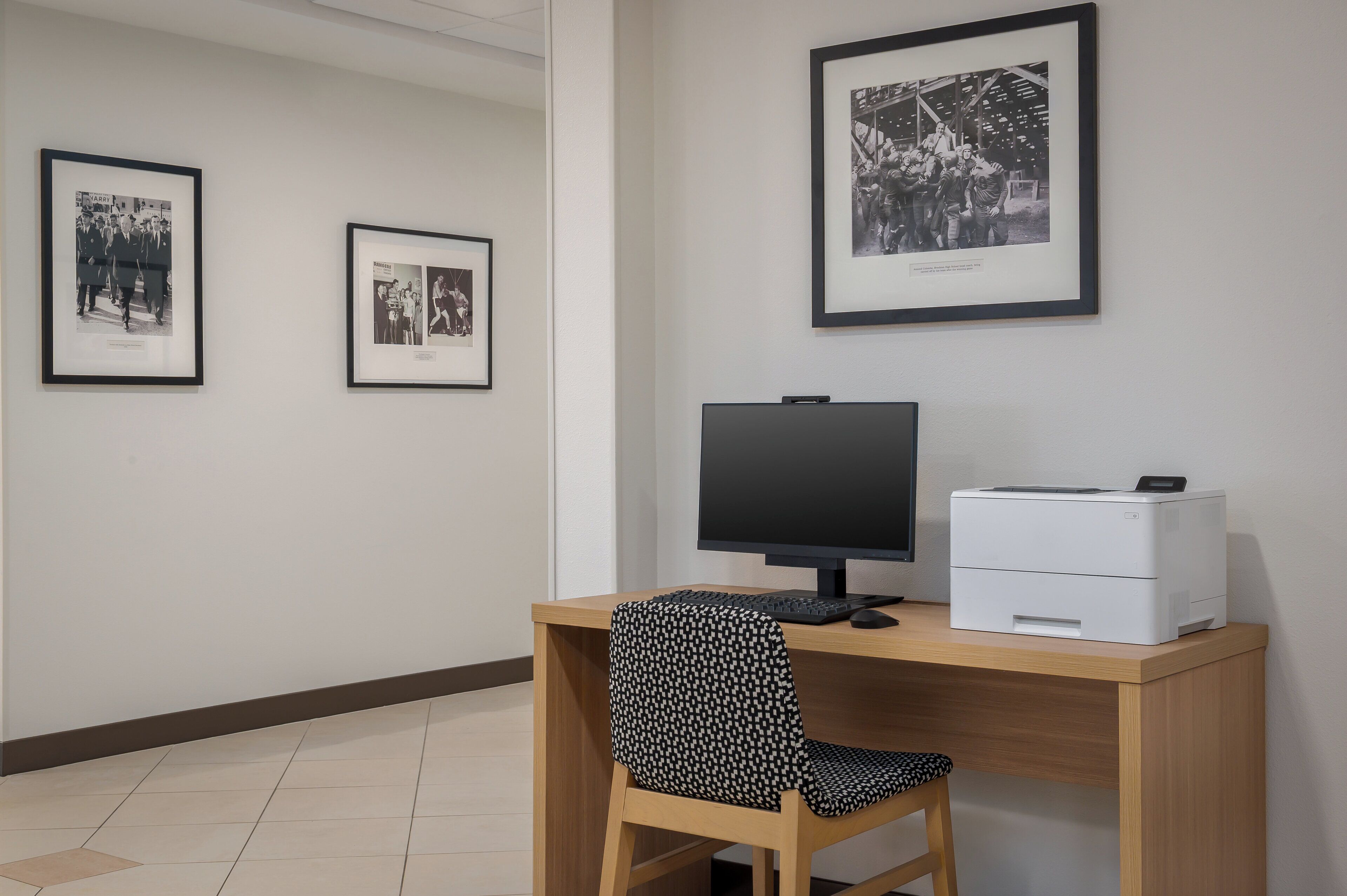 Een werkplek met een bureau, computer, printer en twee ingelijste foto's aan de muur.
