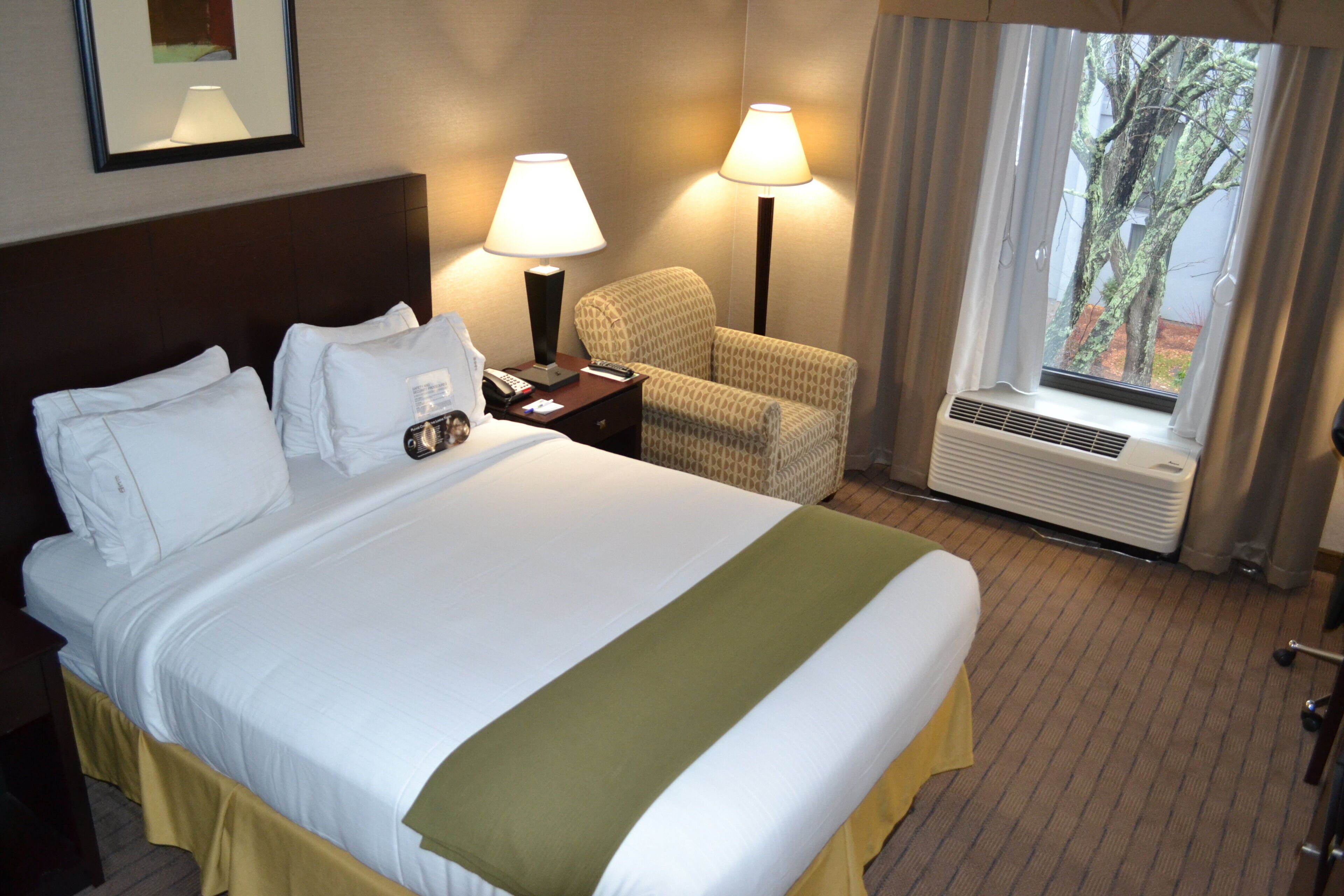 Una camera d'albergo con un letto, una sedia, un comodino con una lampada e una finestra con tende.
