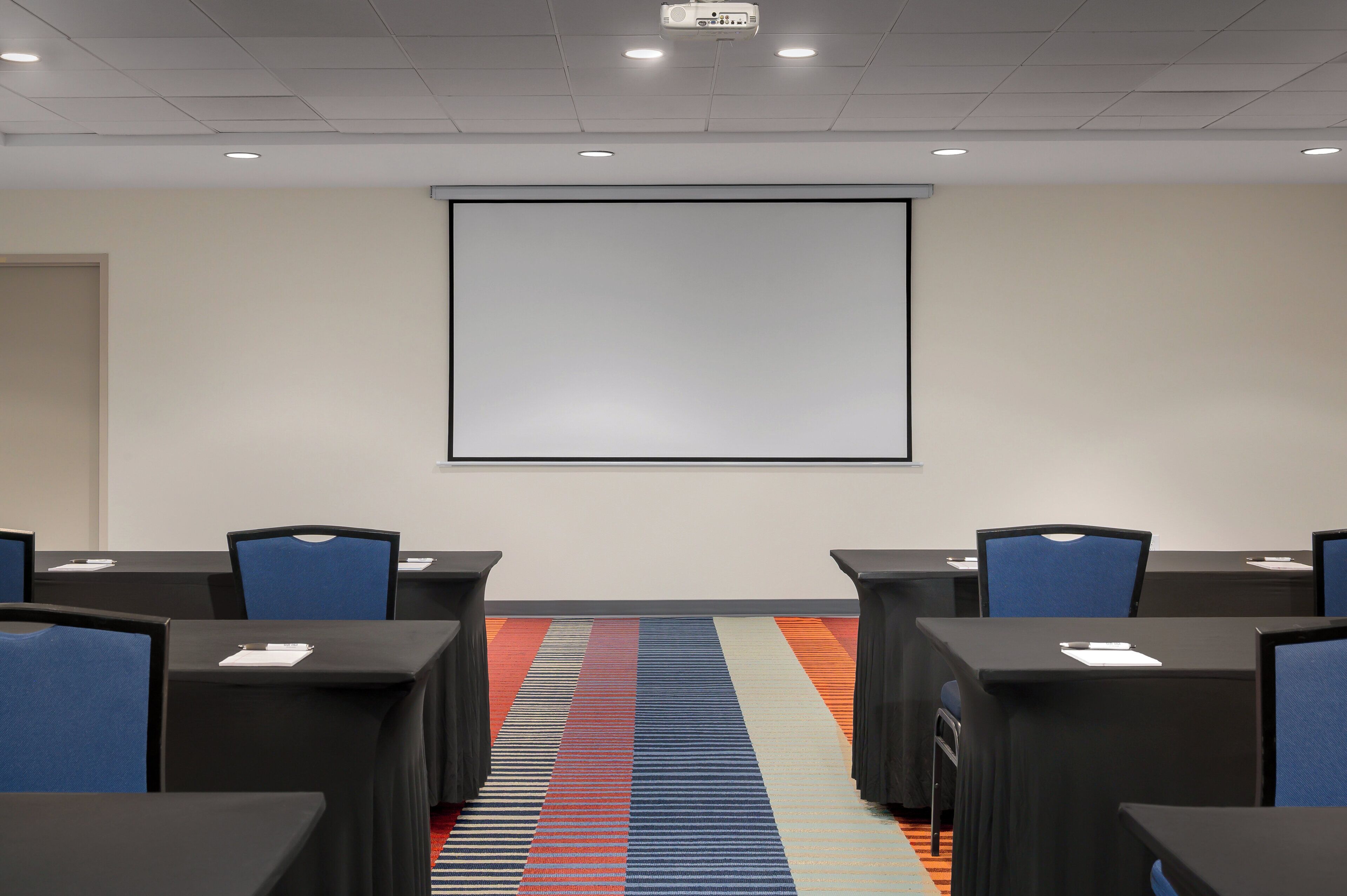 A conference room with a projector screen, tables, chairs, and a carpeted floor.