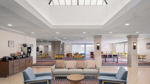 A modern hotel lobby with a sectional sofa, a coffee station, and a dining area with colorful chairs.