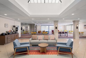 A modern hotel lobby with a sectional sofa, a coffee station, and a dining area with colorful chairs.