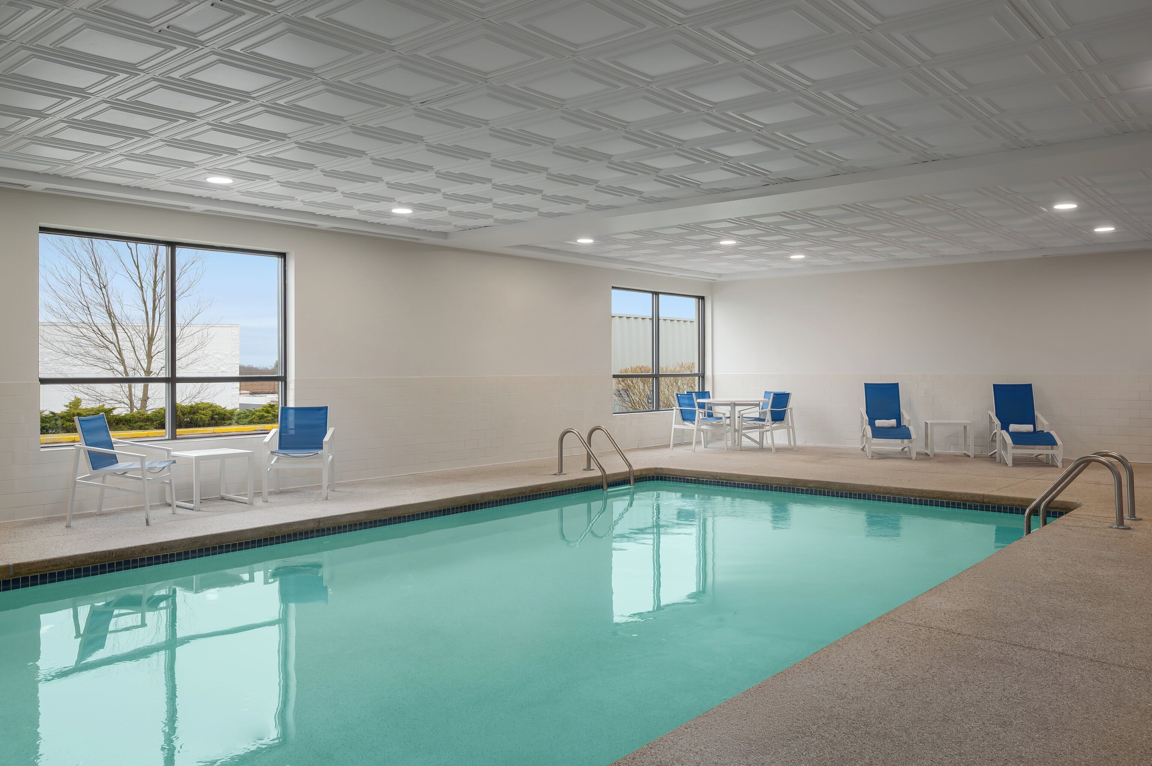 An indoor swimming pool with clear blue water, white poolside chairs, and a tiled ceiling.