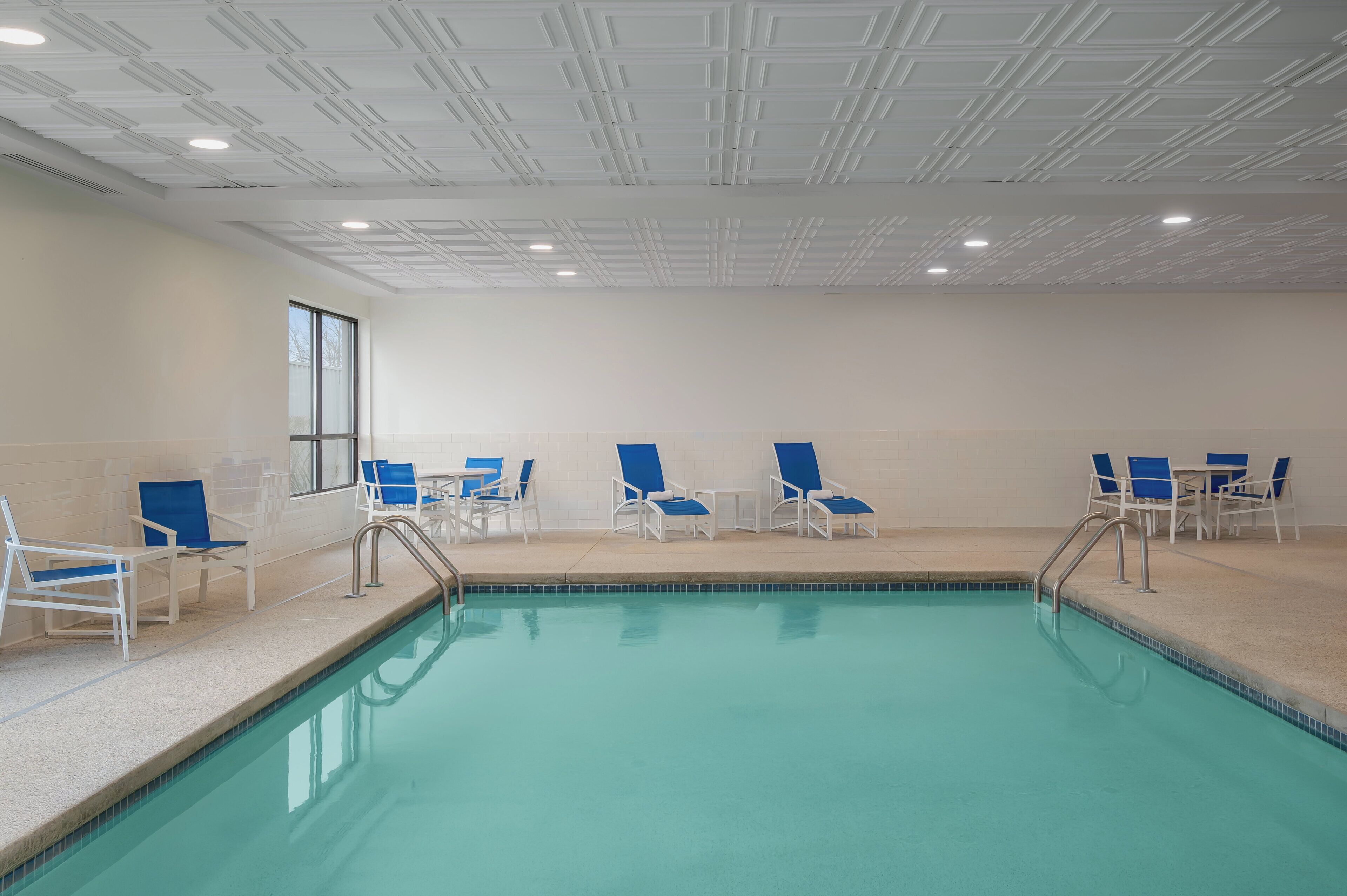 An indoor swimming pool with clear blue water, surrounded by white lounge chairs and a tiled floor.