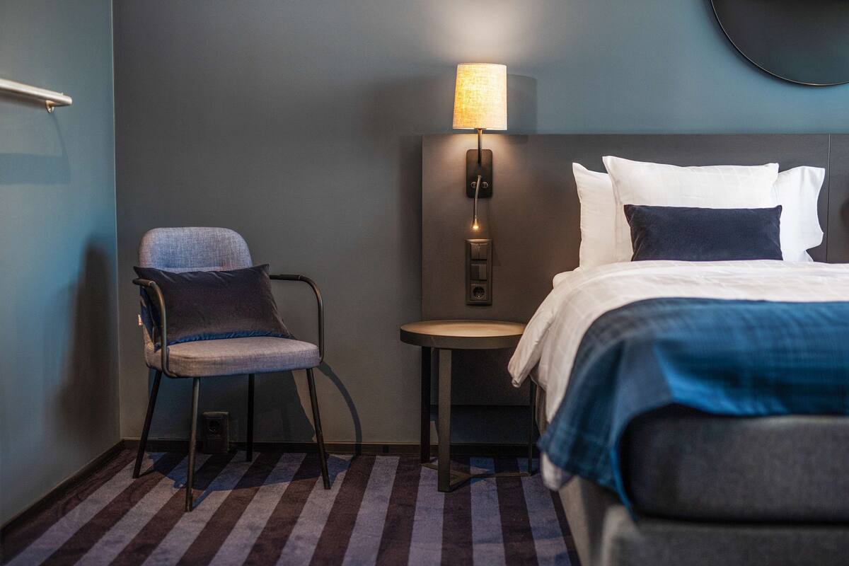 Una habitación de hotel con una cama, una silla, una mesita y una lámpara fijada a la pared.