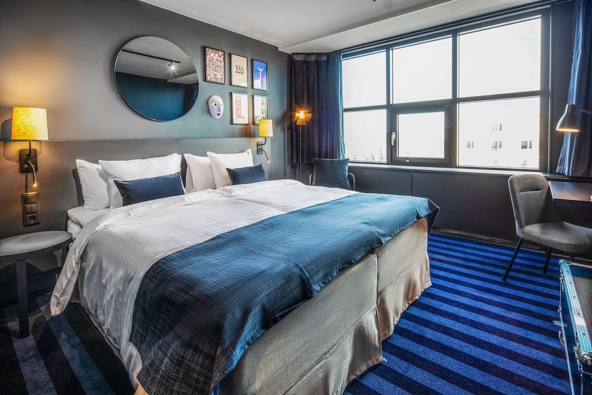 Una habitación de hotel con una cama grande, dos mesitas de noche, una silla, un ventanal con cortinas y decoración en la pared.