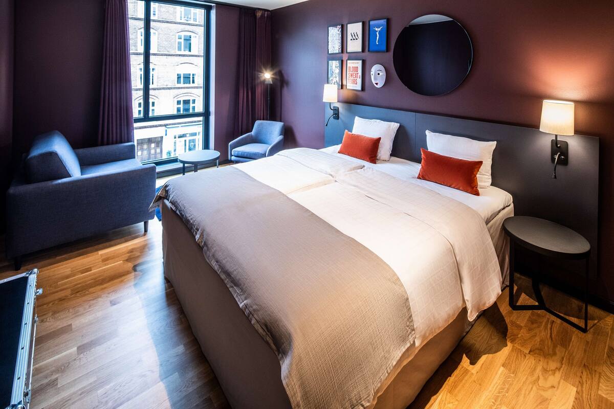 Habitación de hotel con una cama grande, un sofá, un escritorio y vistas de edificios a través de la ventana.