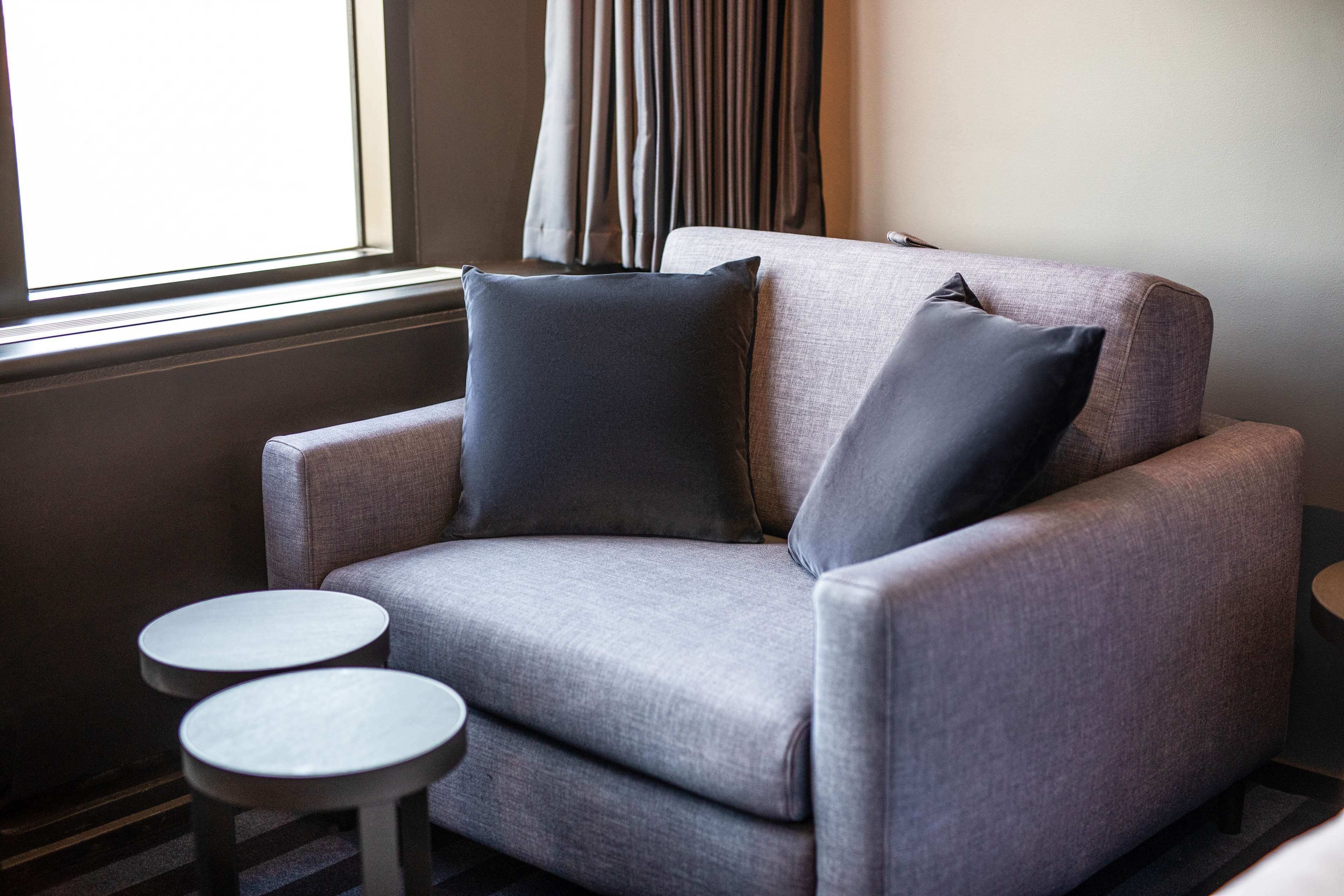 Un sillón gris con dos cojines oscuros, una mesita redonda y una ventana con cortinas.