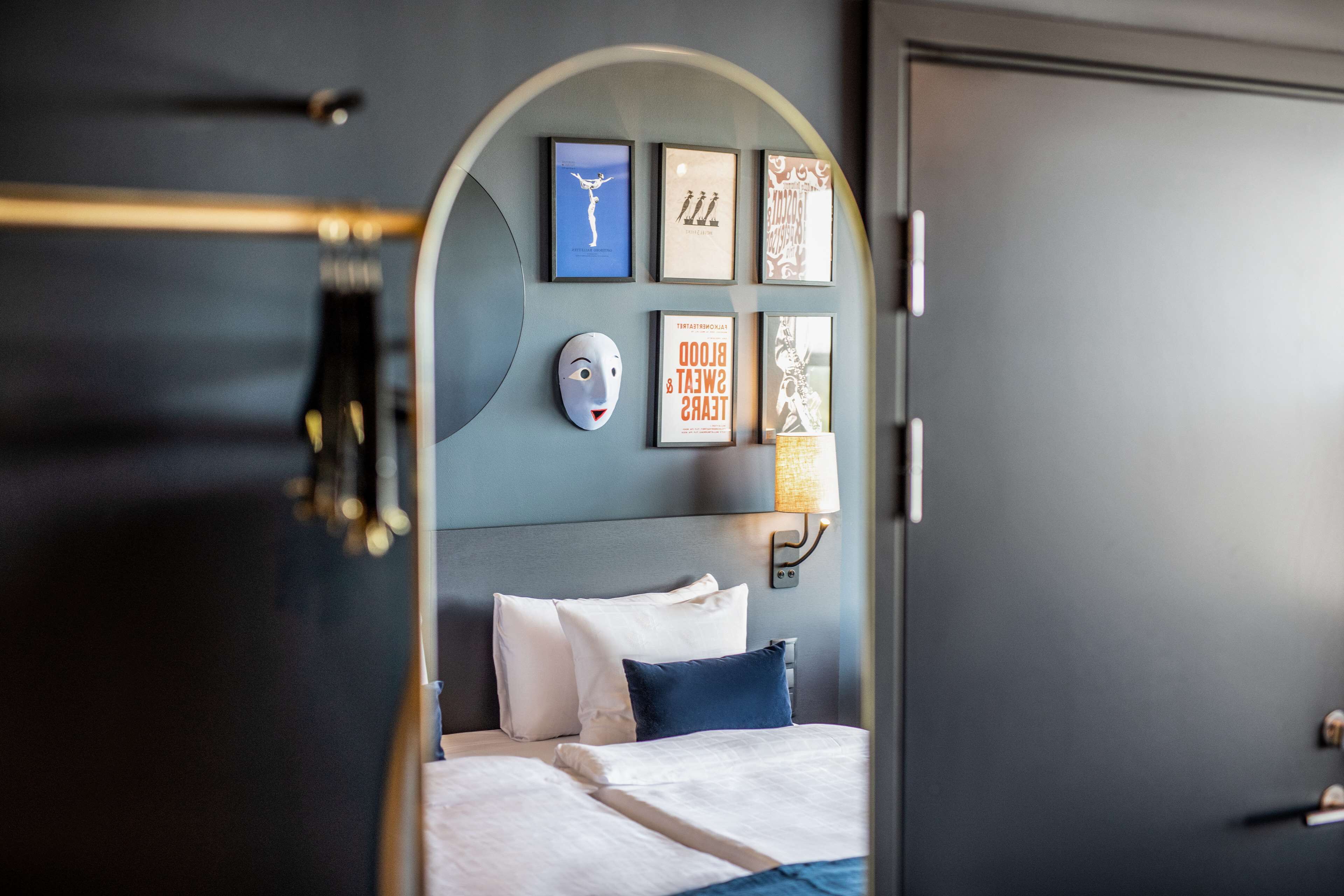 Una camera d'albergo con un letto, una testiera, una lampada a parete e opere d'arte incorniciate.