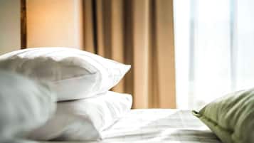 Una cama bien hecha con ropa de cama blanca y un almohadón verde, una lámpara y una ventana con cortinas al fondo.